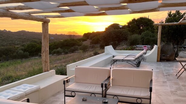 An outdoor patio with a pergola, cushioned seating, and a view of the sunset over a hilly landscape.