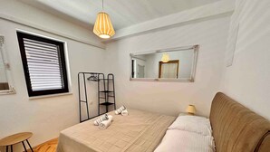 A bedroom with a bed, a window with blinds, a mirror, a hanging light fixture, and a small table. - Holiday apartment with balcony and sea view (Crikvenica)