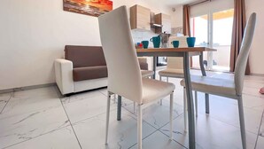 A modern living room with a sofa, a dining table with chairs, and a kitchen area with cabinets and a coffee machine.