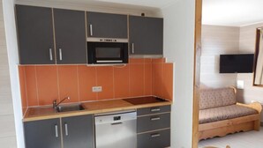 A compact kitchen with grey cabinets, a built-in oven, and a dishwasher. There's a wooden dining table and a sofa in the adjacent living area.