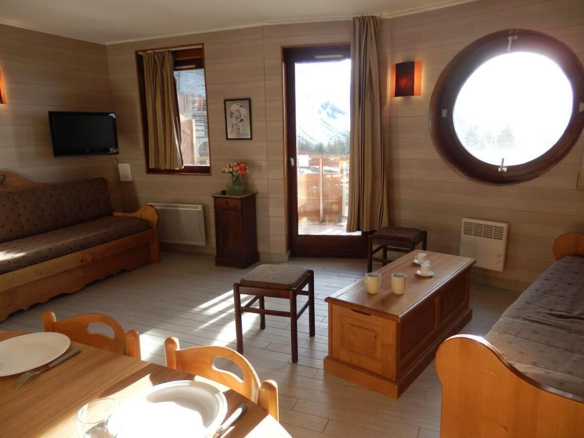A cozy room with a wooden floor, a sofa, a dining table with chairs, a small wooden desk, and a round window with a view of mountains.