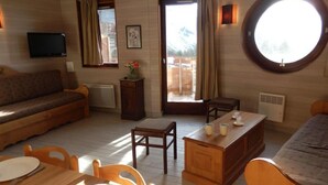A cozy room with a wooden floor, a sofa, a dining table with chairs, a small wooden desk, and a round window with a view of mountains.