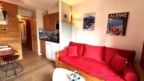 A cozy room with a red sofa, a wooden dining table with chairs, a kitchen area, and a wall with two framed pictures.
