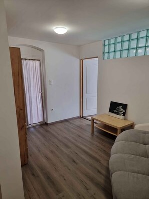Een kamer met houten vloer, een houten deur, een witte muur, een kleine tafel met een fotolijstje en een grijze bank.