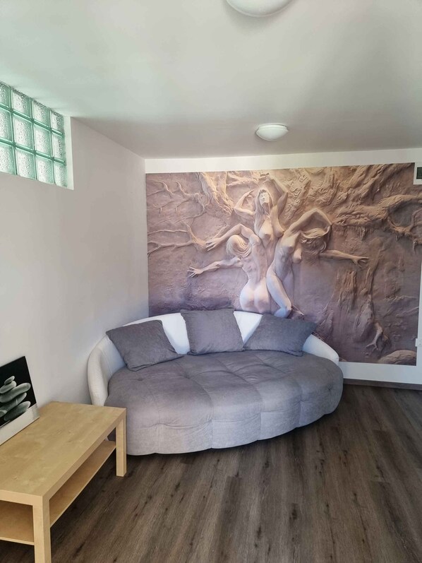 Een moderne kamer met een gebogen grijze bank, een klein houten tafeltje en een groot wanddecoratie van een boom en figuren.
