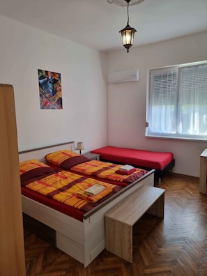 Een slaapkamer met een bed, een bankje, een raam met gordijnen, een airconditioning en een schilderij aan de muur.