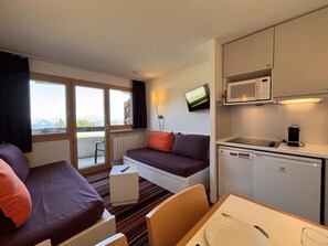 Quarto de hotel com sofá, mesa pequena, cozinha compacta e televisão.