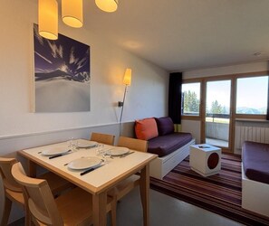 A modern room with a dining table set for four, a sofa with cushions, a small white side table, and a large window with a view.