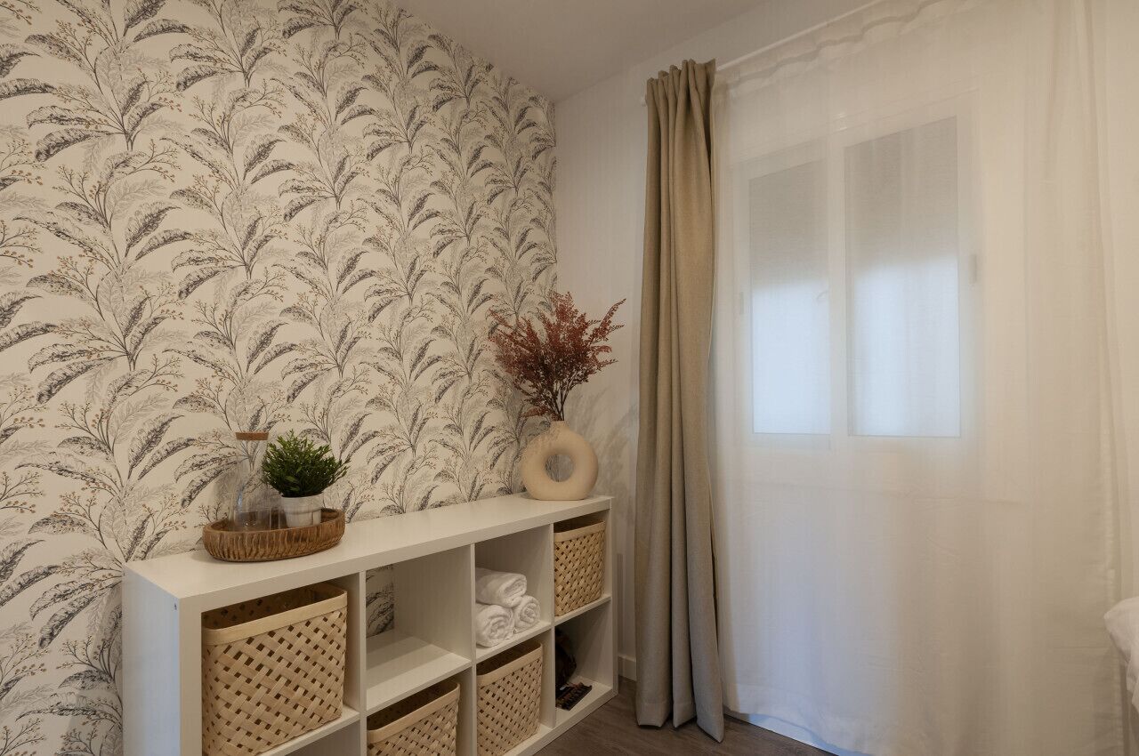 Une pièce avec un papier peint à motifs floraux, une console blanche agrémentée de paniers et une fenêtre avec des rideaux.