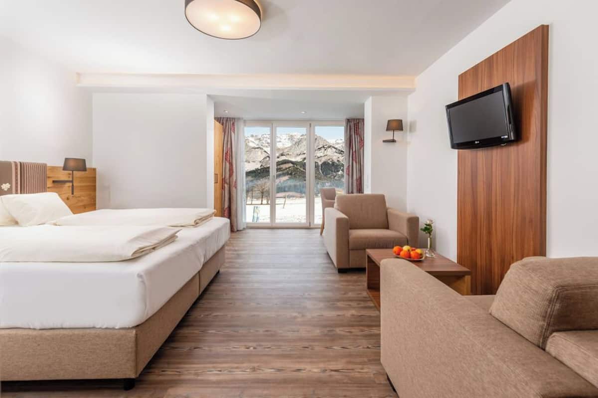 Ein Hotelzimmer mit Bett, Sofa, Wandfernseher und Blick auf Berge durch das Fenster.