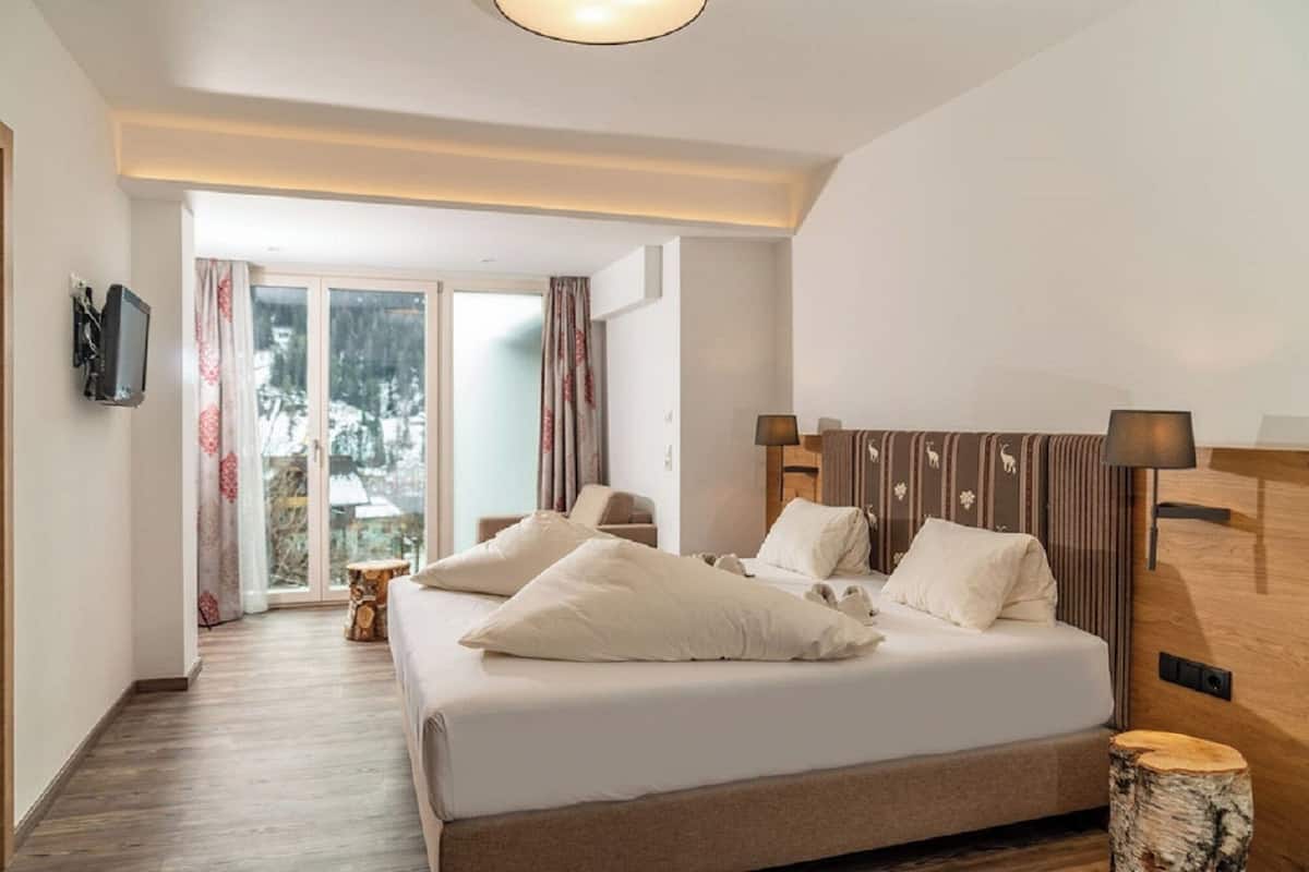 Ein Schlafzimmer mit einem großen Bett, einem hölzernen Kopfteil, einem Nachttisch, einer Lampe und Blick auf eine verschneite Landschaft durch eine Glastür.
