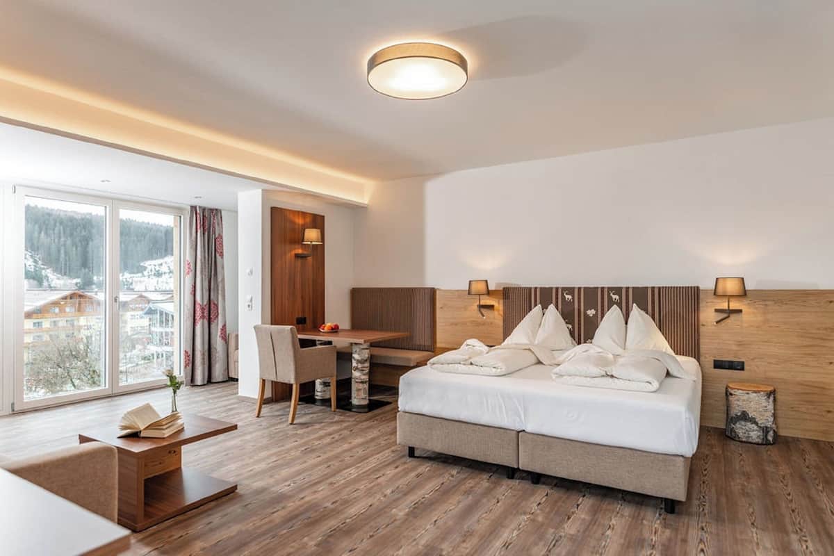 Ein modernes Hotelzimmer mit einem großen Bett, einem Schreibtisch und einem Sessel. Ein Fenster bietet Blick auf Gebäude und Bäume.