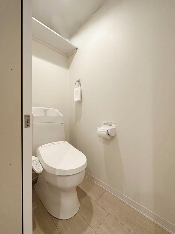 A small, clean bathroom with a toilet, a trash bin, and a toilet paper holder on the wall. - F Style (Hiroshima)