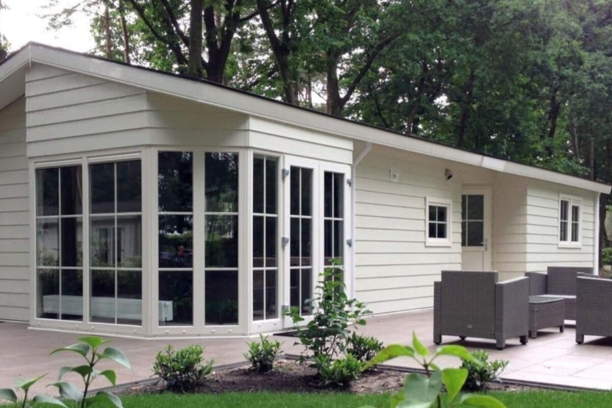 Una casa moderna con un amplio ventanal de vidrio, un porche cubierto con mobiliario exterior y un jardín cuidado.