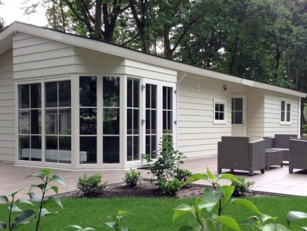 A modern house with a large glass bay window, a covered patio with outdoor furniture, and a well-maintained garden.