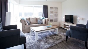 Un salon moderne comprenant un canapé, deux fauteuils, une table basse, un téléviseur et des œuvres d’art accrochées au mur.
