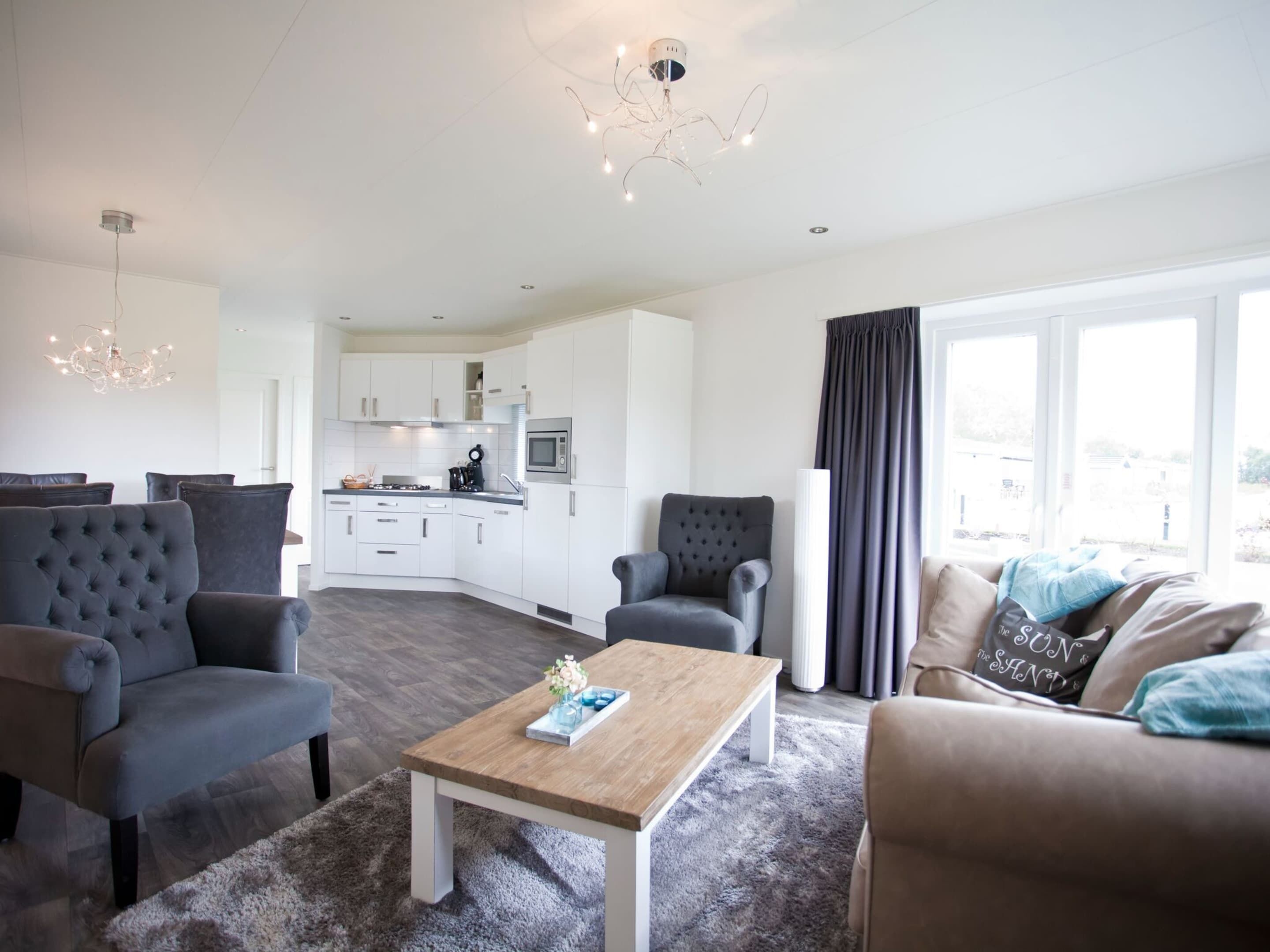 A modern living room with a sofa, armchairs, a coffee table, and a view of the kitchen in the background.