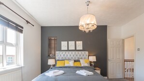 A bedroom with a grey bed, a chandelier, a window with blinds, and a door.
