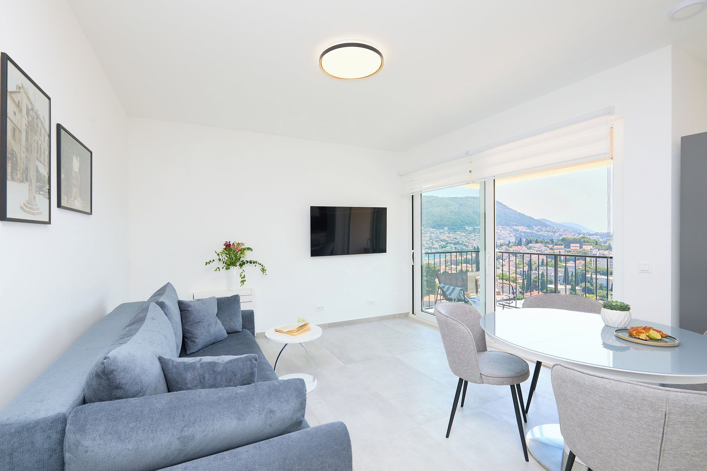 A modern living room with a grey sofa, a small white table, a TV mounted on the wall, and a balcony with a view of the city and mountains.