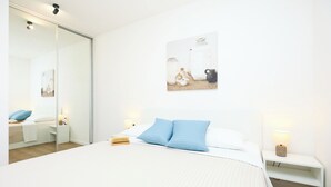 A modern bedroom with a large bed, a bedside table with a lamp, a wall-mounted painting, and a sliding glass door leading to another room.