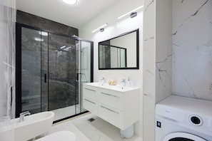 Une salle de bain moderne dotée d’une douche à parois de verre, d’un meuble-lavabo blanc avec un pare-feu en marbre et d’un lave-linge encastré.
