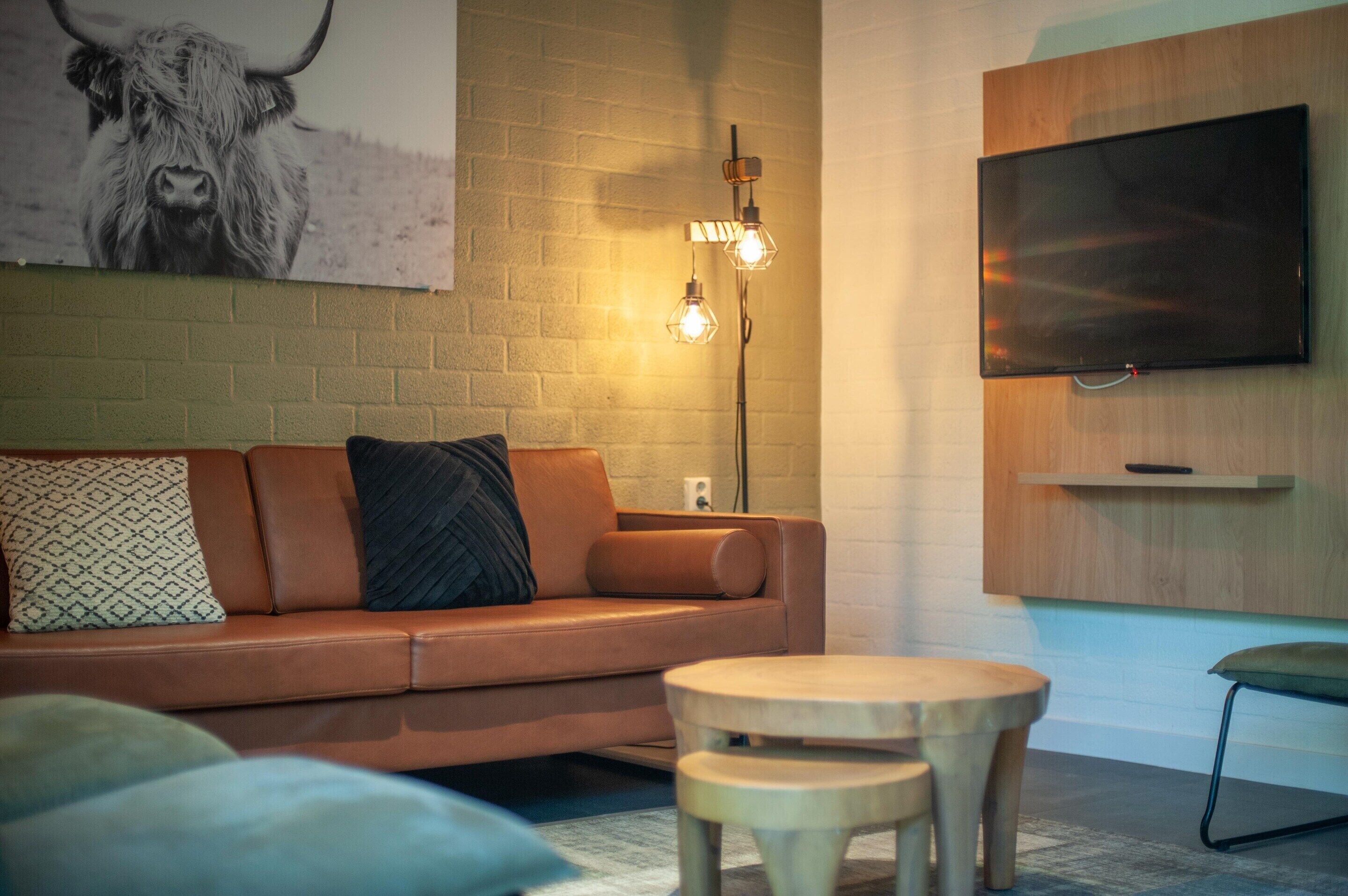 A modern living room with a brown leather sofa, two wooden side tables, a mounted flat-screen TV, and a wall-mounted light fixture.