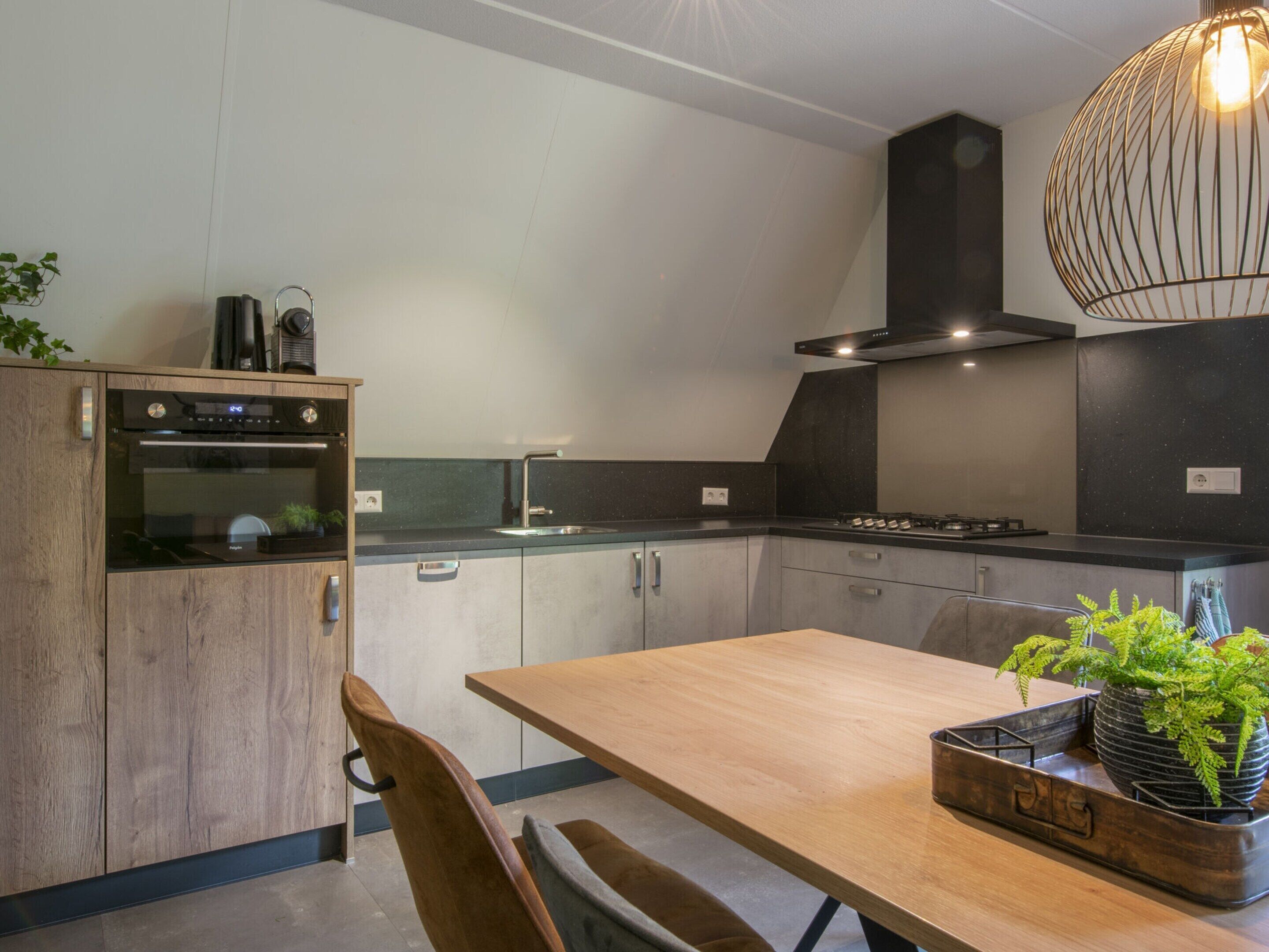 A modern kitchen with a wooden table, two chairs, a built-in oven, a sink, and a hanging light fixture.