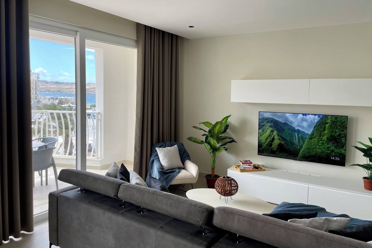 A modern living room with a grey sofa, a TV mounted on the wall, a potted plant, and a view of the sea through large windows.