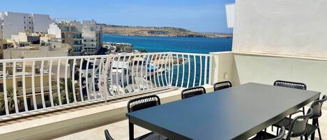 A balcony with a table and chairs, overlooking a coastal area with buildings and a clear blue sea.