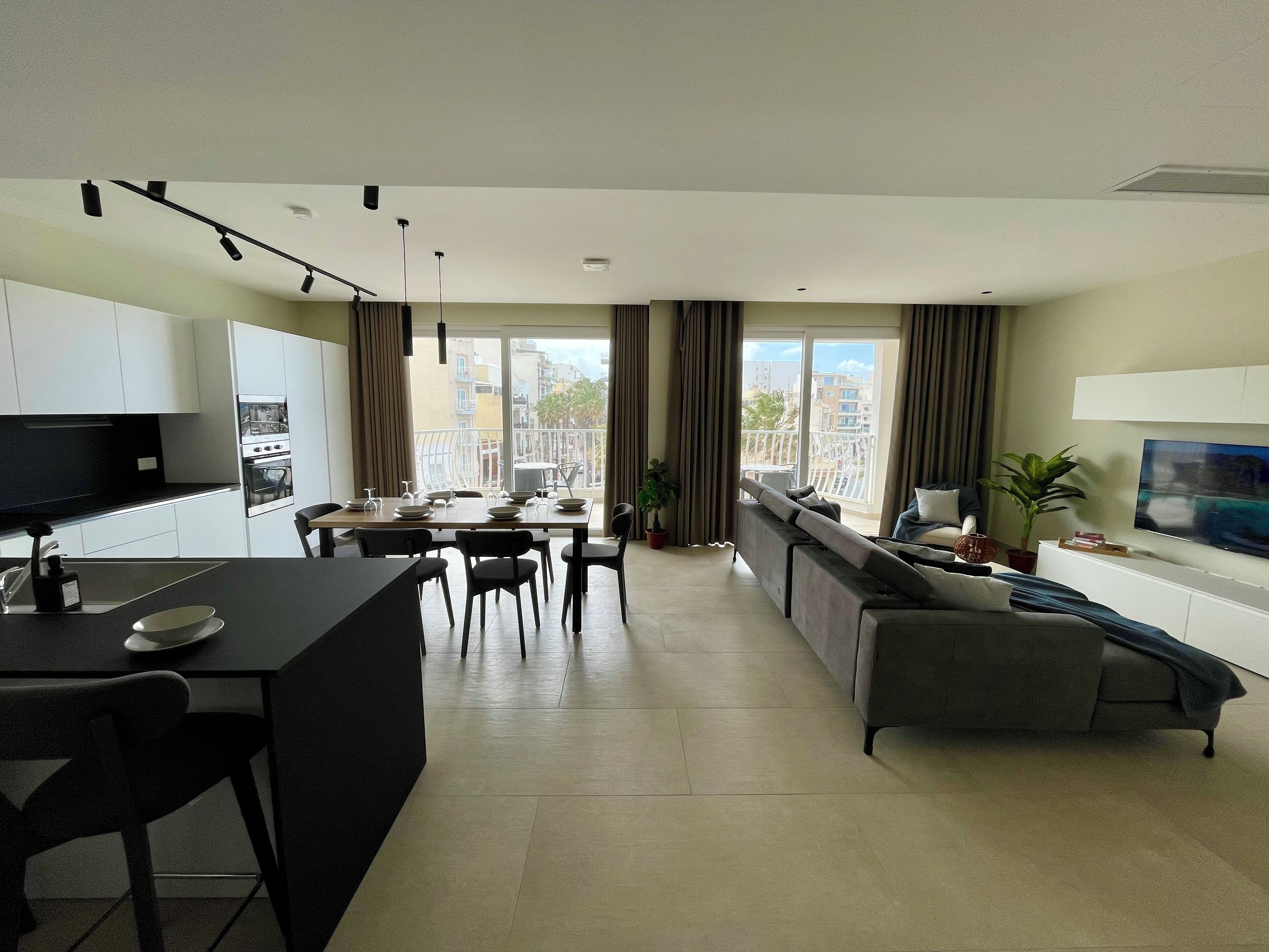 A modern apartment with a kitchen, dining area, and living room. It features a large window with a city view, a flat-screen TV, and a sofa.