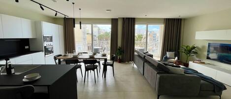 A modern apartment with a kitchen, dining area, and living room. It features a large window with a city view, a flat-screen TV, and a sofa.