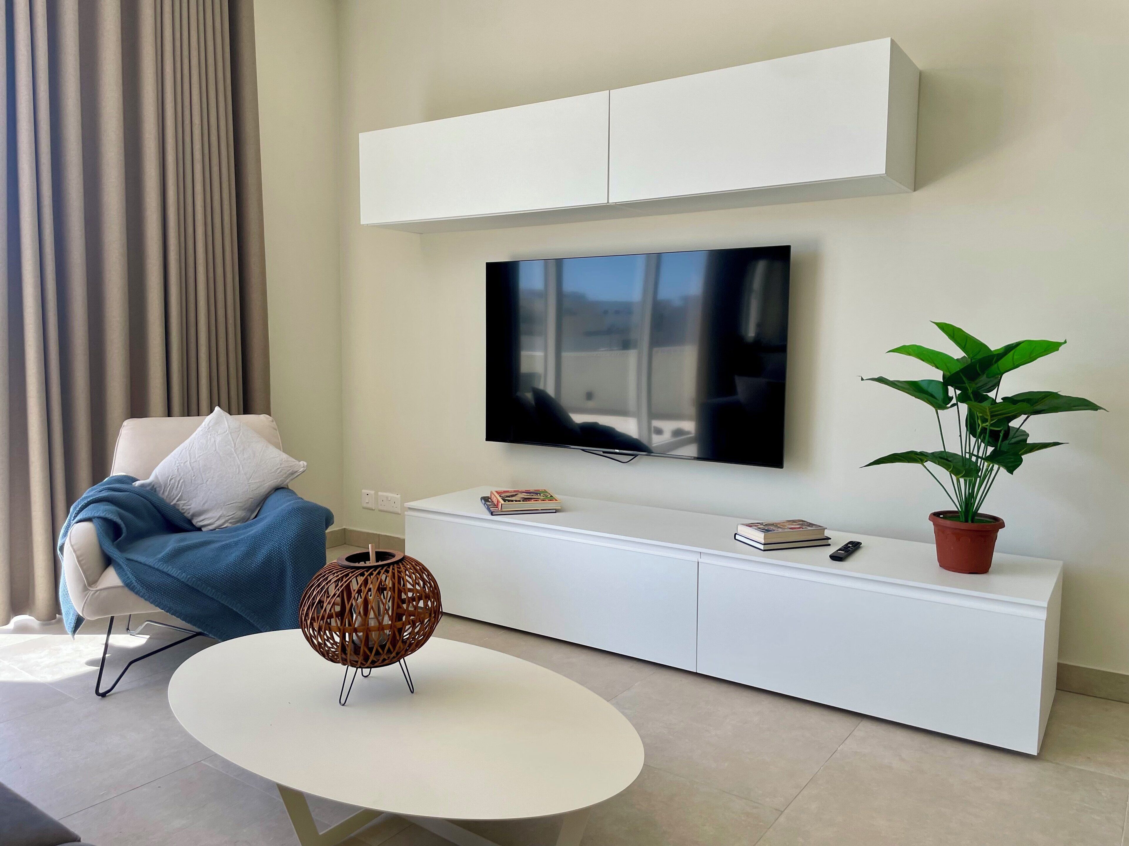 A modern living room with a flat-screen TV mounted on a white cabinet, a comfortable sofa with a blue throw blanket, a round coffee table, a potted plant, and a decorative wicker lamp.