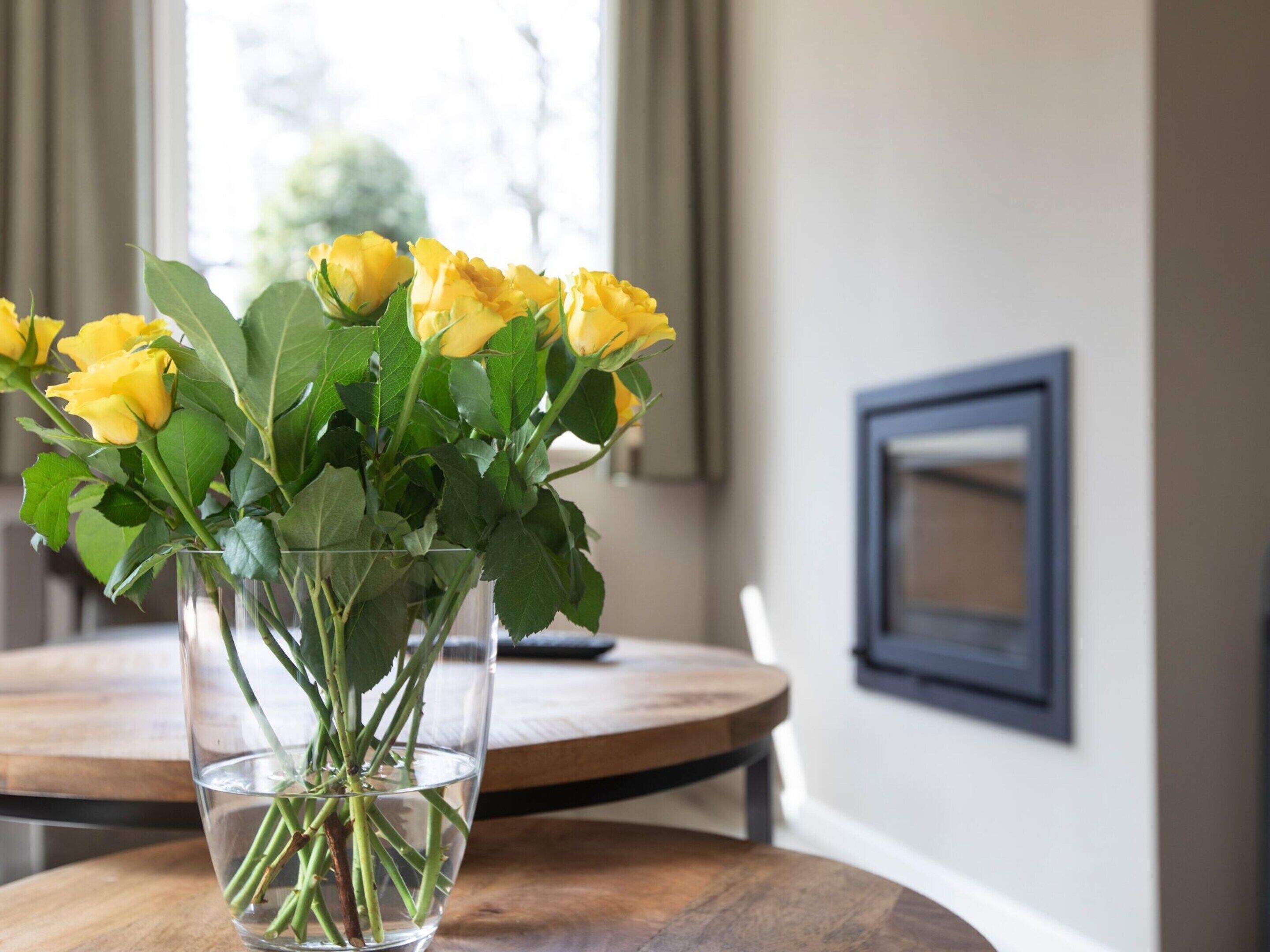 A clear glass vase with yellow roses on a wooden table in a room with a window and a framed picture on the wall.
