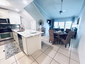 A kitchen with white cabinets, a built-in microwave, and a dining area with wicker chairs and a table. - 🏖️Last minute dates available ☀️JULY☀️Don’t miss out on this Beautiful condo!! (Miramar Beach)
