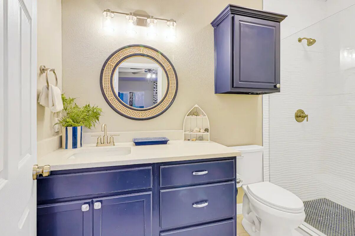 A bathroom with a white sink, dark blue cabinets, a round mirror with a gold frame, and a white toilet.