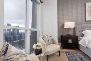 A modern bedroom with a large window, a bedside table with a lamp, a grey upholstered bed, and a chair with a vase of flowers.