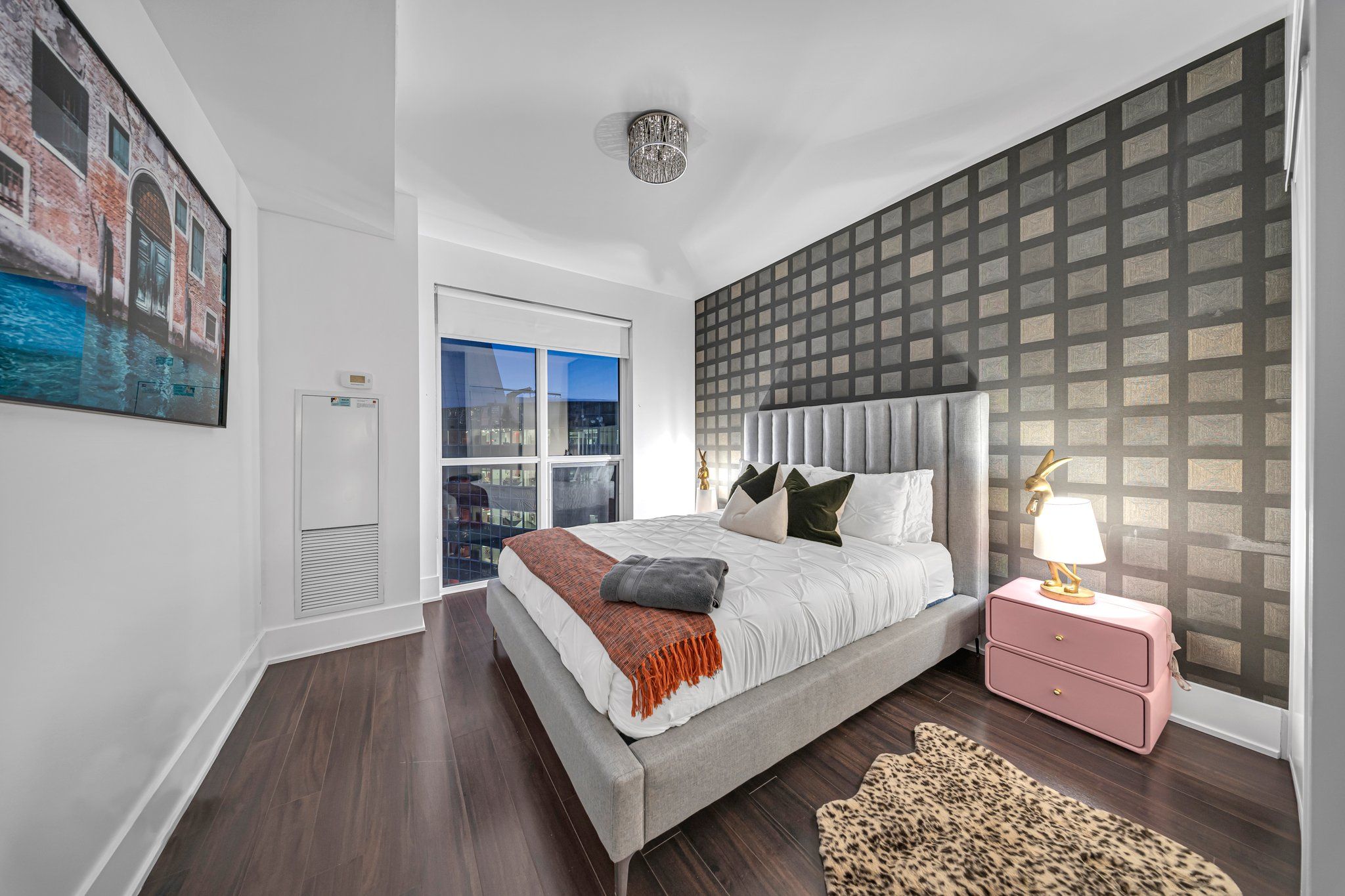 A modern bedroom with a large bed, a nightstand, a lamp, and a view of the city through a window.