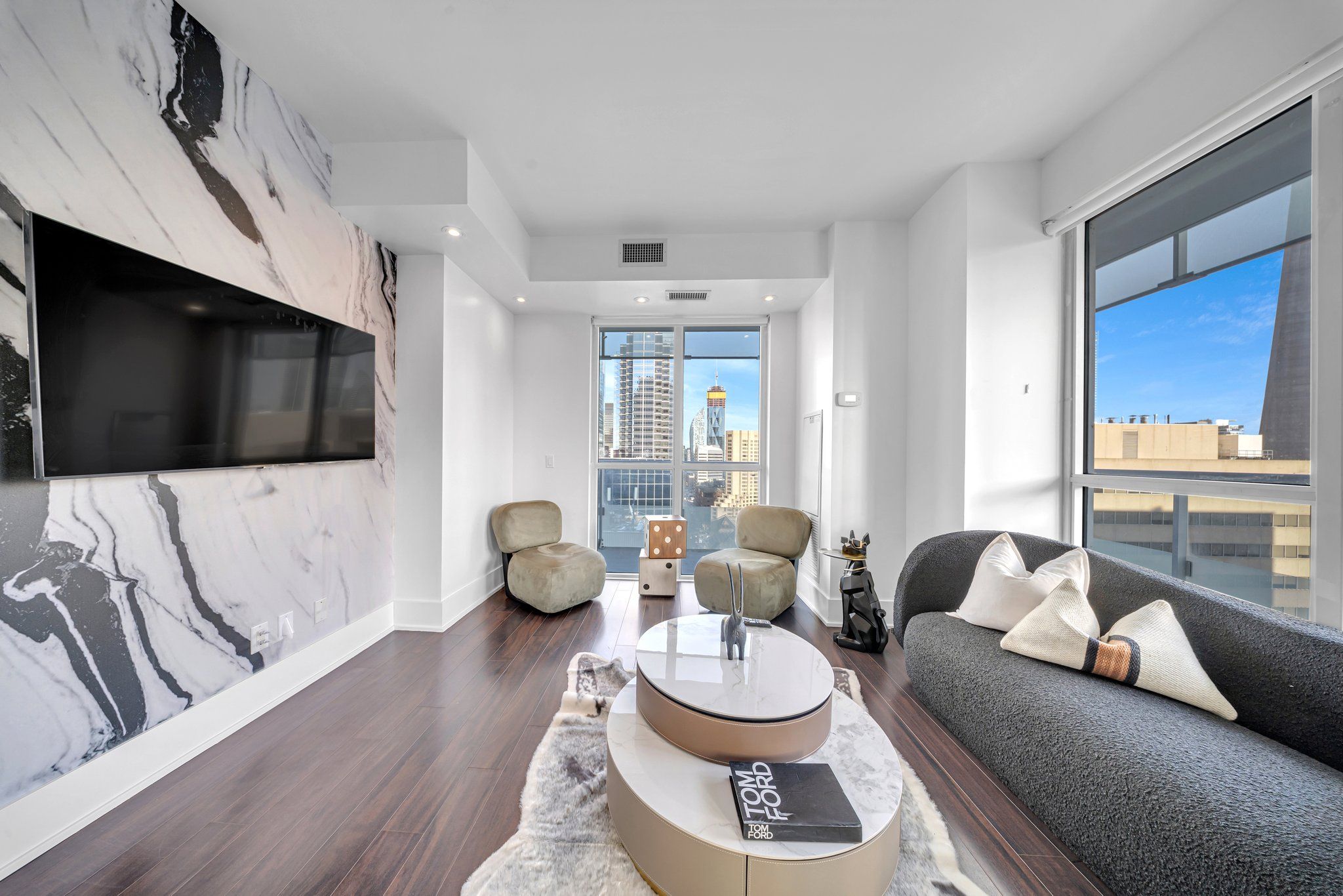 A modern living room with a large TV, comfortable seating, a coffee table, and a view of the cityscape.