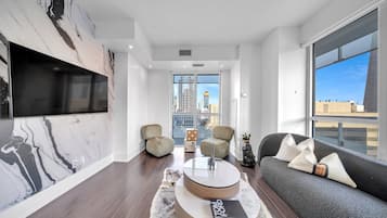 A modern living room with a large TV, comfortable seating, a coffee table, and a view of the cityscape.