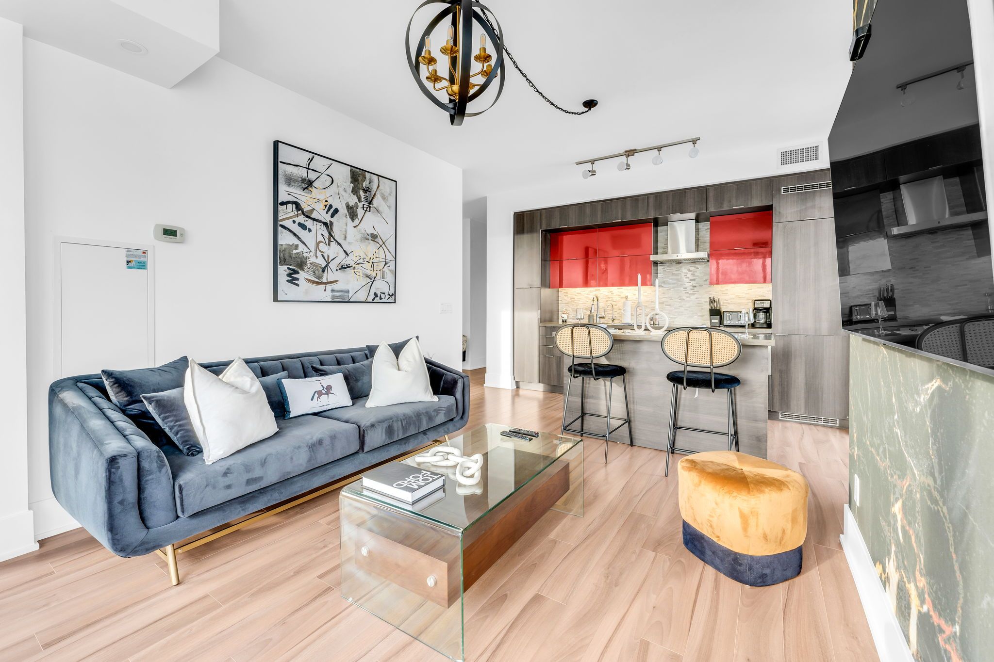 A modern living room with a sofa, coffee table, and ottoman. There is a kitchen area with red cabinets and a dining set.