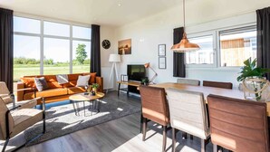 A modern living room with a sofa, coffee table, dining table, and chairs. There is a TV, lamp, and artwork on the wall.