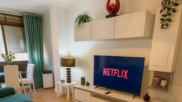 Una sala de estar moderna con un televisor de pantalla plana mostrando 'NETFLIX', una mesa de centro, sofás color turquesa y un jarrón rojo decorativo.