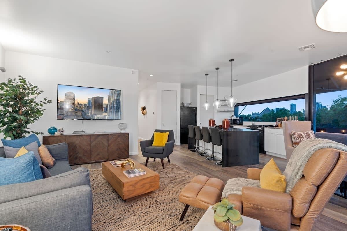 A modern living space with a sofa, armchair, ottoman, and a coffee table. There is a cityscape painting on the wall, a plant, and a kitchen area visible through an open door.