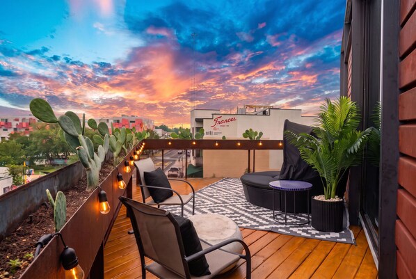 A rooftop terrace with a view of the city skyline, featuring a modern outdoor seating area with chairs and a table, surrounded by potted plants and a string of lights. - 2BR Designer Condo East Downtown Austin (Austin)