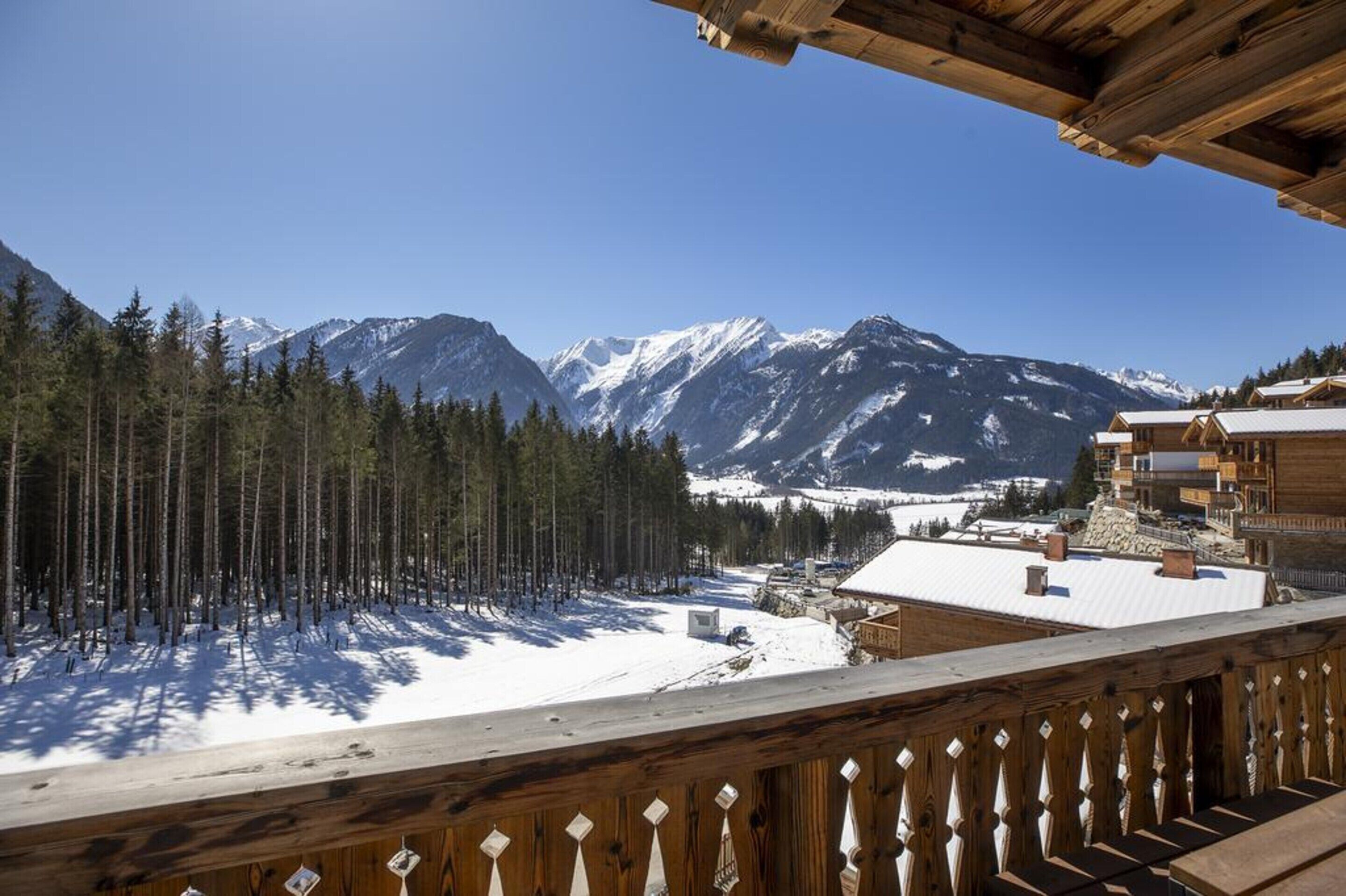 Un chalet de montagne doté d’un balcon en bois offrant une vue sur un paysage enneigé et des montagnes aux sommets couverts de neige.