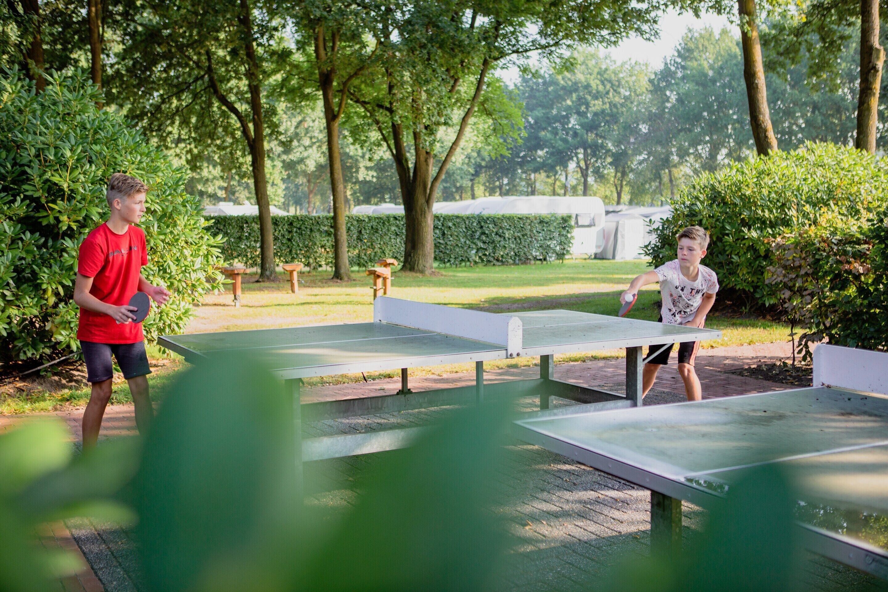 Two people playing table tennis on an outdoor court surrounded by trees and greenery.