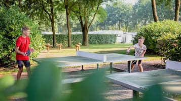 Two people playing table tennis on an outdoor court surrounded by trees and greenery.