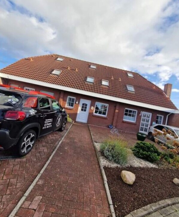 Une maison à deux étages, avec un toit recouvert de tuiles rouges, une allée pavée en briques et un petit jardin agrémenté de rochers et d’arbustes.