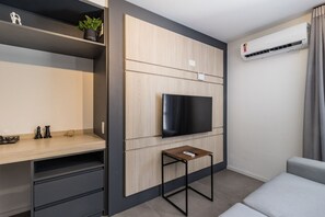 A modern living room with a flat-screen TV mounted on a wooden panel wall, a small wooden table with a remote, and a grey sofa. - Duque by My Way (Porto Alegre)