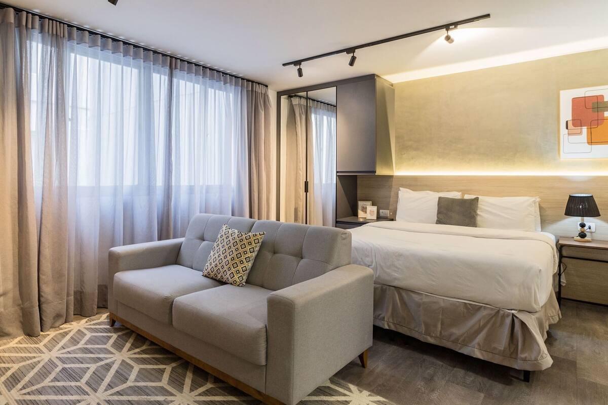 Quarto de hotel com sofá, cama, mesa de cabeceira, luminária e iluminação embutida na parede.